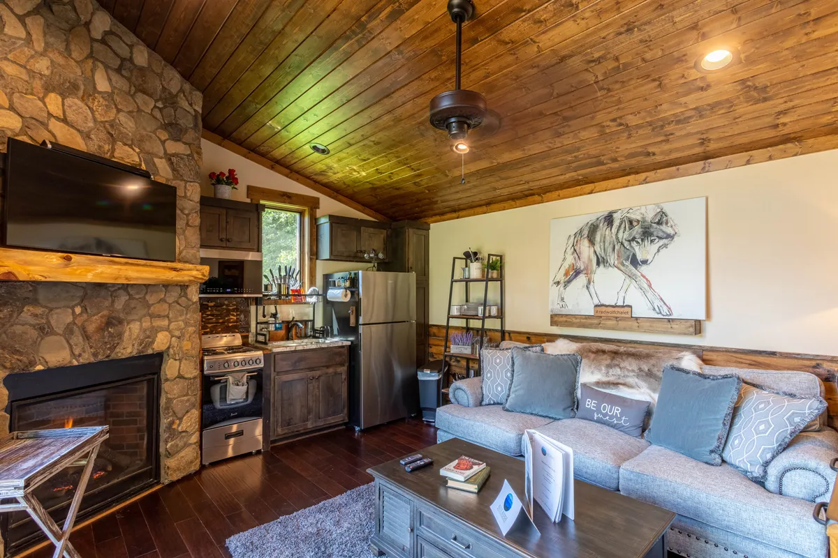 A welcoming living area featuring a stone fireplace as the focal point, flanked by a comfortable sofa and accented with warm lighting. The open-plan layout allows a glimpse into the adjoining kitchen area, while artwork above the fireplace adds character and a touch of intimacy.