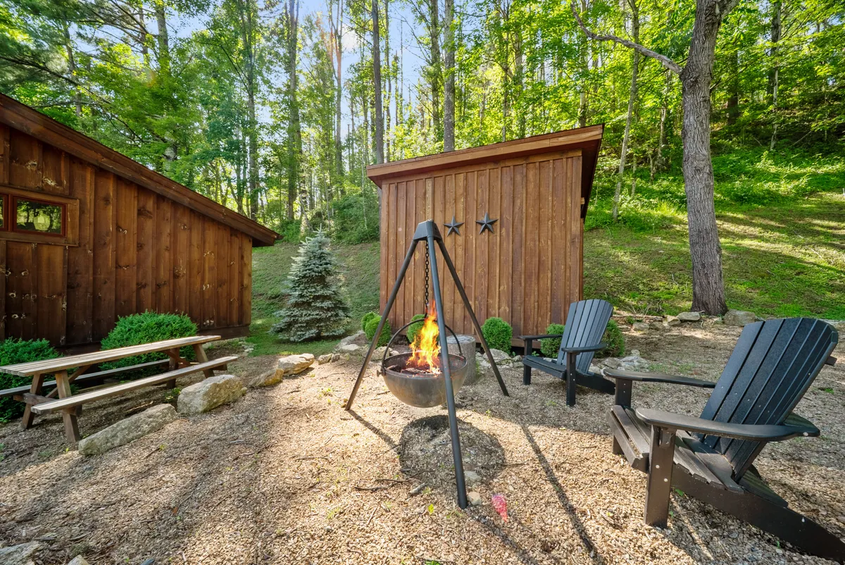 A cozy fire pit encircled by Adirondack chairs, with rustic wooden cabins in the background and a picnic table nearby. The scene is set at dusk, with the glow of the fire and the night sky beginning to appear.