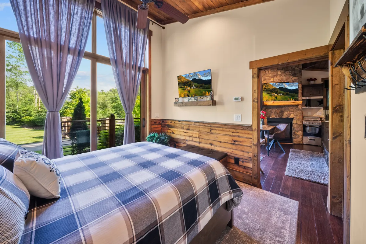 This inviting bedroom scene features a plaid bedspread, large windows with flowing curtains, wooden wall paneling, and curated artwork. The setting exudes warmth and coziness, with natural light enhancing the tranquil mood.