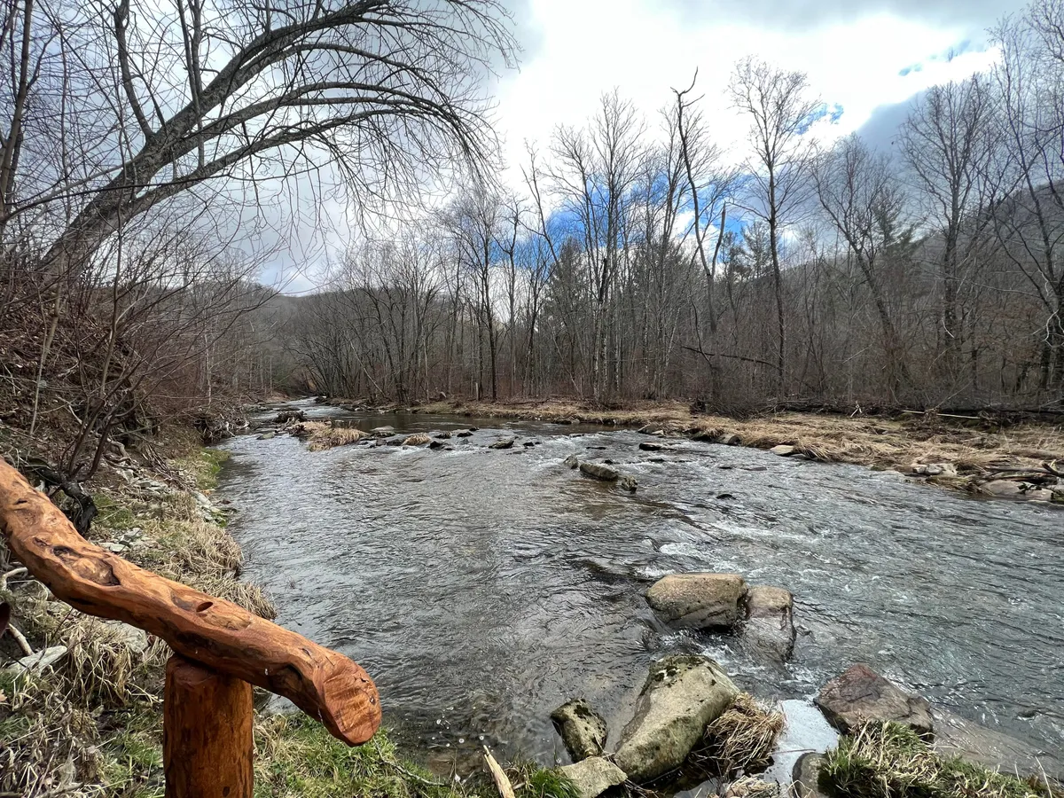 A tranquil view of a gently flowing river bordered by rock formations, trees, and a wooden railing, evoking a peaceful natural setting ideal for fishing or relaxing by the water.