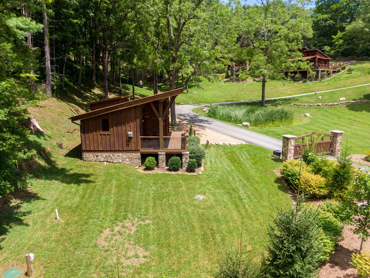 A picturesque view of the wooden cabin set amidst a lush, landscaped lawn, surrounded by tall trees and natural greenery. The stone pathway leads to a gated entrance, creating a welcoming and secure environment for guests and their dogs. The outdoor space appears expansive and inviting, ideal for pets to roam and for owners to relax in nature.