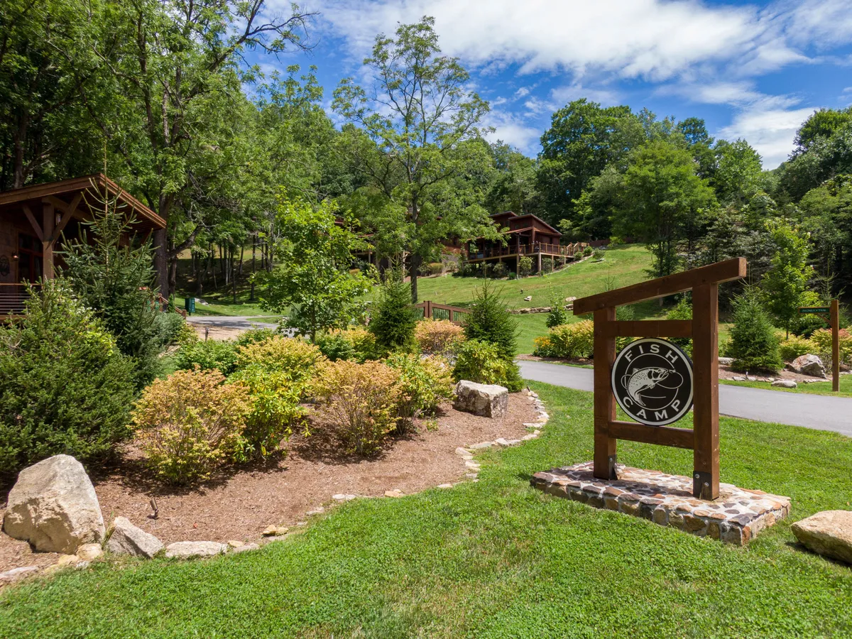 A welcoming wooden sign with the 'Fish Camp' logo stands amidst lush greenery, with charming scenic cabins visible in the background. The image captures the essence of an outdoor-focused community, inviting guests to engage in activities like fishing, hiking, and campfire gatherings.