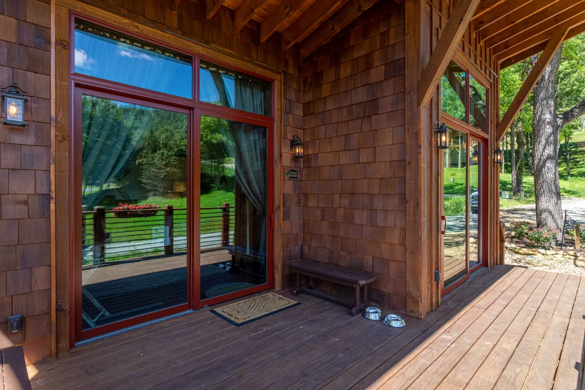 The chalet’s wooden deck features large glass doors opening to an inviting outdoor seating area, surrounded by lush greenery. The warm glow of sunset or morning light suggests moments of relaxation—ideal for shared coffees, intimate conversations, or enjoying the mountain ambiance.