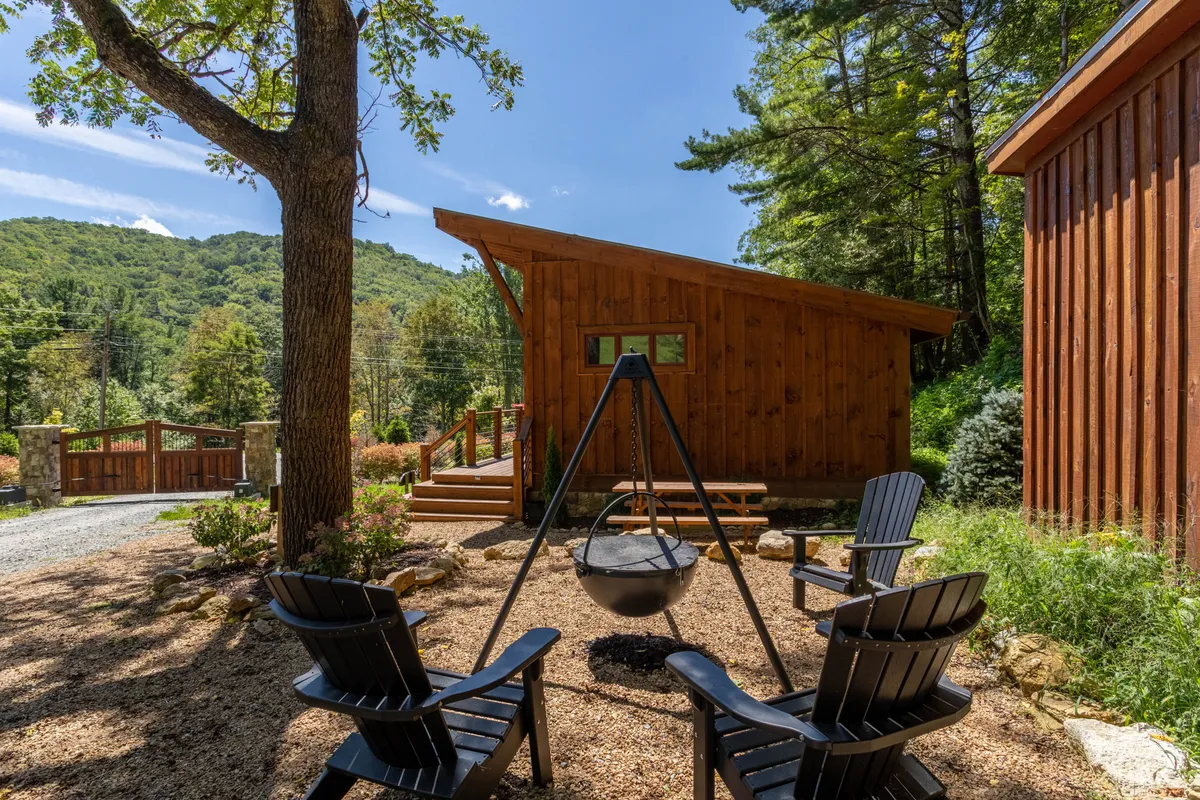 This image showcases a charming wooden cabin set against a mountain backdrop, with Adirondack chairs arranged around a Cowboy Cauldron-style fire pit. The scene is tranquil, suggesting private moments by the fire and inviting stargazing. The surrounding nature and relaxed seating create an atmosphere perfect for couples seeking romantic seclusion.