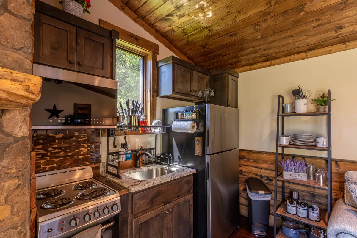 A warmly lit kitchen interior featuring sleek cabinets, a modern sink, a stove, a refrigerator, and a window that lets in natural light. The area is compact but thoughtfully organized, with utensils and countertop appliances visible, conveying a sense of functionality and coziness.