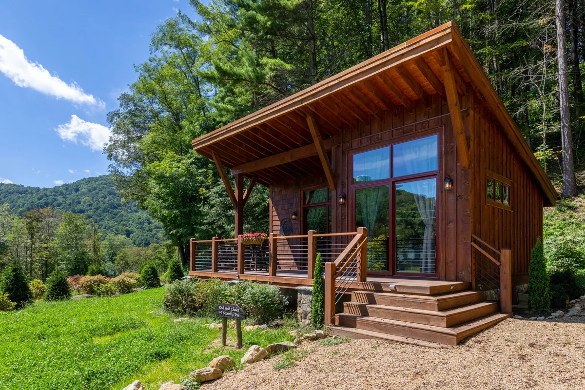 The image showcases a picturesque wooden cabin nestled amidst lush greenery, with a spacious deck overlooking mountain views. Large windows invite natural light, while the cozy deck is perfect for relaxing and taking in the tranquil surroundings. The serene mountain backdrop sets a peaceful tone, ideal for couples seeking a romantic retreat in nature.